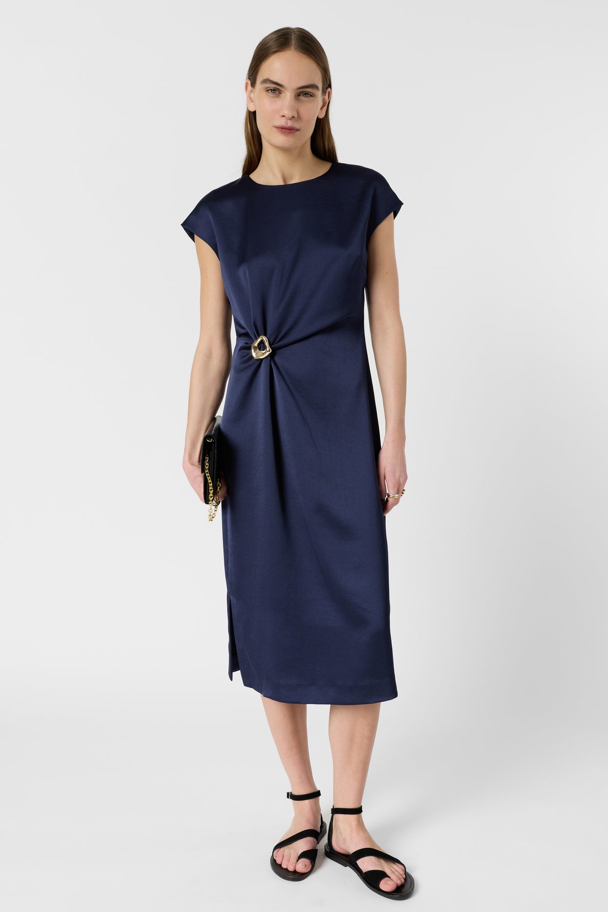 Woman wearing a robe mi-longue bleu marine with a side buckle detail, black sandals, and holding a black clutch, standing against a plain white background.