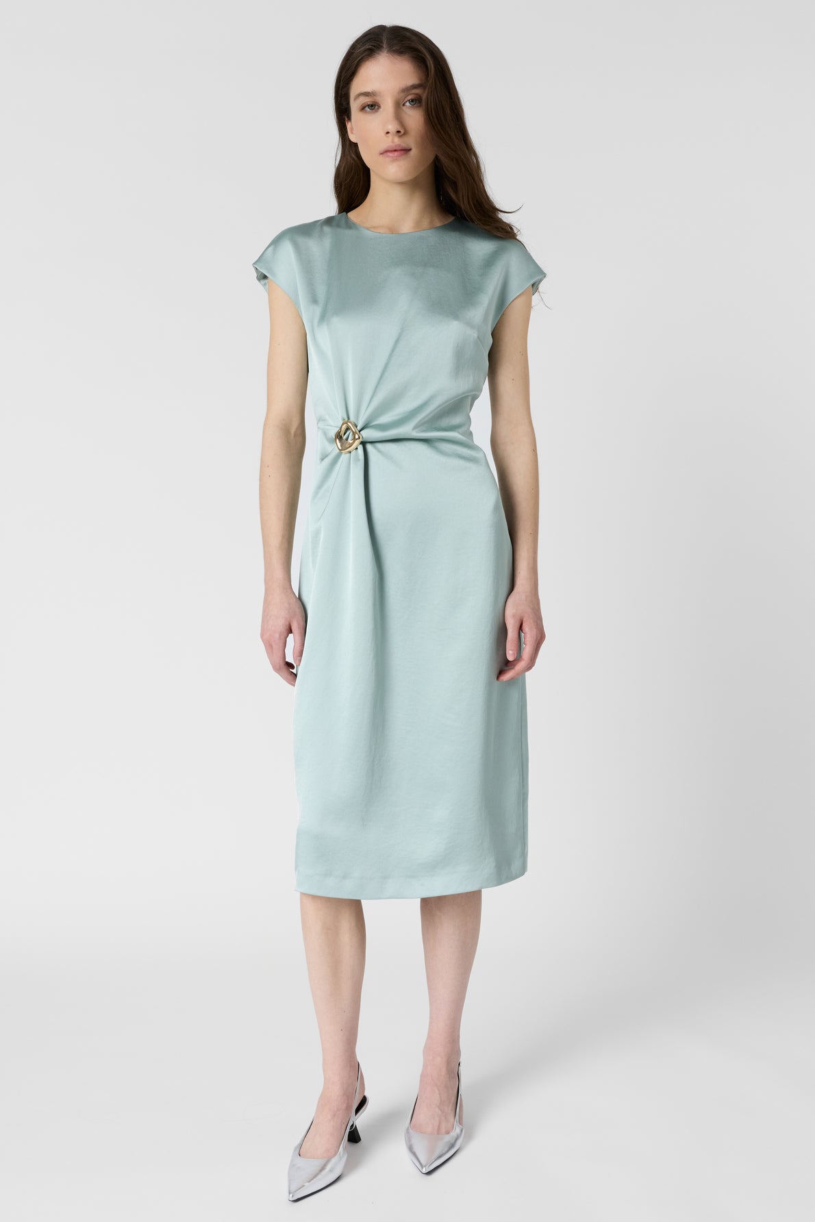 A woman stands facing forward, wearing a knee-length, light blue dress with cap sleeves and a decorative knot at the waist, paired with pointed silver heels. The background is plain white.