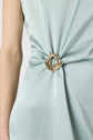 Close-up of a person wearing a light blue dress gathered at the waist with a metallic, abstract-shaped brooch. Only the left arm and torso are visible against a plain background.