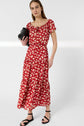 A person wearing a red floral maxi dress and black sandals, holding a black clutch bag, stands against a plain light background.