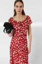 A person wearing a red dress with white floral patterns holds a black handbag and looks at the camera with a neutral expression against a plain background.