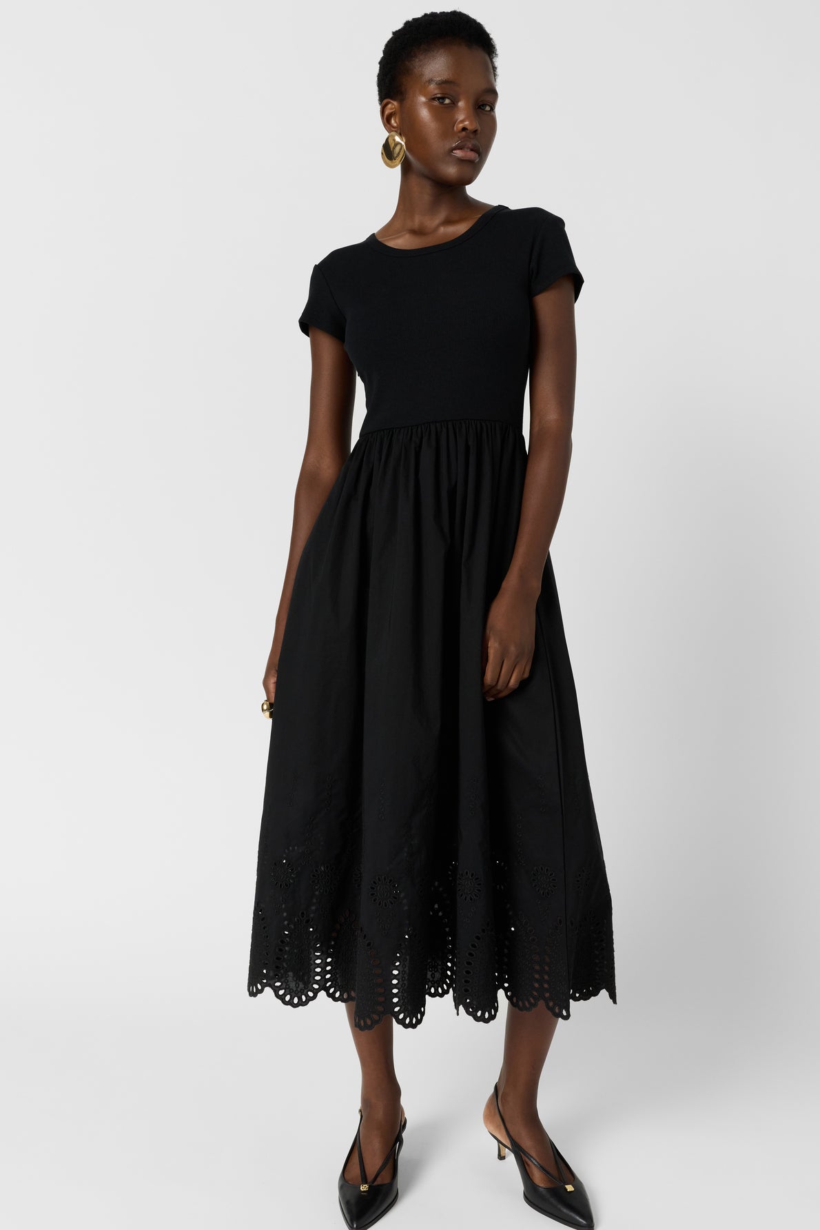 A woman wearing a short-sleeve black dress with a fitted top and flared, eyelet-hem skirt stands against a plain background, accessorized with gold hoop earrings and black pointed heels.