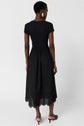 Woman wearing a black, short-sleeve, fitted top with a high-waisted, eyelet-hem midi skirt, standing barefoot and facing away against a plain white background.