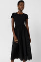 A woman wearing a short-sleeve black midi dress with a fitted top, a gathered skirt, and eyelet details at the hem. She has short hair, gold hoop earrings, and gold rings, standing against a plain background.