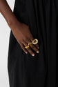 A person wearing a black dress has their hand resting against their hip, displaying several large, bold gold rings on their fingers.
