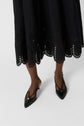 A person wearing a black, laser-cut midi skirt and black pointed-toe slingback heels stands against a white background. Only the lower legs and feet are visible.