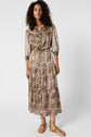 A woman stands wearing a patterned, button-front midi dress with three-quarter sleeves and a floral and paisley print. She has curly hair and is wearing tan strappy sandals, posing against a plain white background.