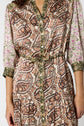A woman wearing a patterned dress with paisley and floral designs in brown, green, and pink tones. The dress has green-trimmed edges, buttons down the front, and a matching belt at the waist.