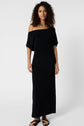 Woman with curly hair wearing an élégante robe longue in jersey noir, off-the-shoulder and ankle-length, paired with black sandals, stands against a plain white background, looking at the camera.