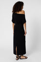 A woman with curly hair stands facing away, wearing an off-shoulder robe longue en jersey noir with a back slit and black sandals, exuding pure élégance against a plain white background.