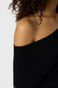 A close-up of a person with curly hair wearing a black off-the-shoulder top, radiating élégance as they reveal their bare shoulder and part of their arm against a plain background.