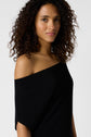 Woman with curly hair wearing an off-the-shoulder black top in jersey noir, standing against a plain white background, looking softly at the camera, exuding effortless élégance.
