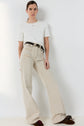 A person stands against a white wall, wearing a white tee-shirt Mavie adorned with a monogramme brodé, light beige high-waisted wide-leg pants with a black belt, and brown loafers with embellishments. Their hair is pulled back and they have a neutral expression.