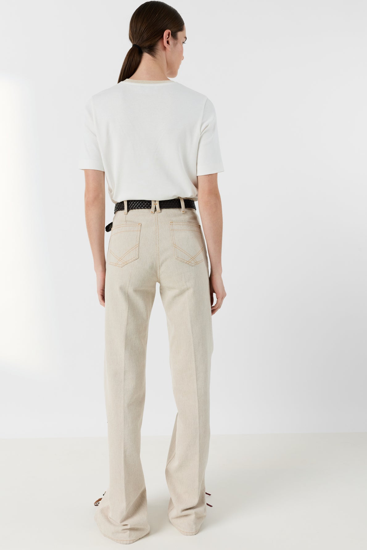 A person with long brown hair in a low ponytail wears a white tee-shirt Mavie with a col doré and subtle monogramme brodé, paired with beige high-waisted pants and a black belt, standing with their back facing the camera against a plain white background.