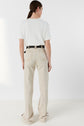 A person with long brown hair in a low ponytail wears a white tee-shirt Mavie with a col doré and subtle monogramme brodé, paired with beige high-waisted pants and a black belt, standing with their back facing the camera against a plain white background.