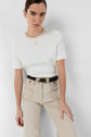 A person with dark hair pulled back wears a crisp white tee-shirt Mavie, featuring a subtle monogramme brodé, and high-waisted beige jeans with a black braided belt, standing against a plain white background.