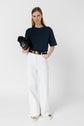 A woman stands against a white background, wearing a navy blue t-shirt, high-waisted white pants, and white sandals. She holds a black clutch and has straight, light brown hair.