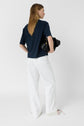 Person with long light brown hair wearing a navy short-sleeve shirt and loose white pants, holding a black bag, standing against a plain white background, facing away from the camera.