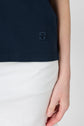 A person wearing a navy blue shirt with a small embroidered logo and white pants is shown from torso to mid-thigh, with their right arm relaxed at their side against a plain background.