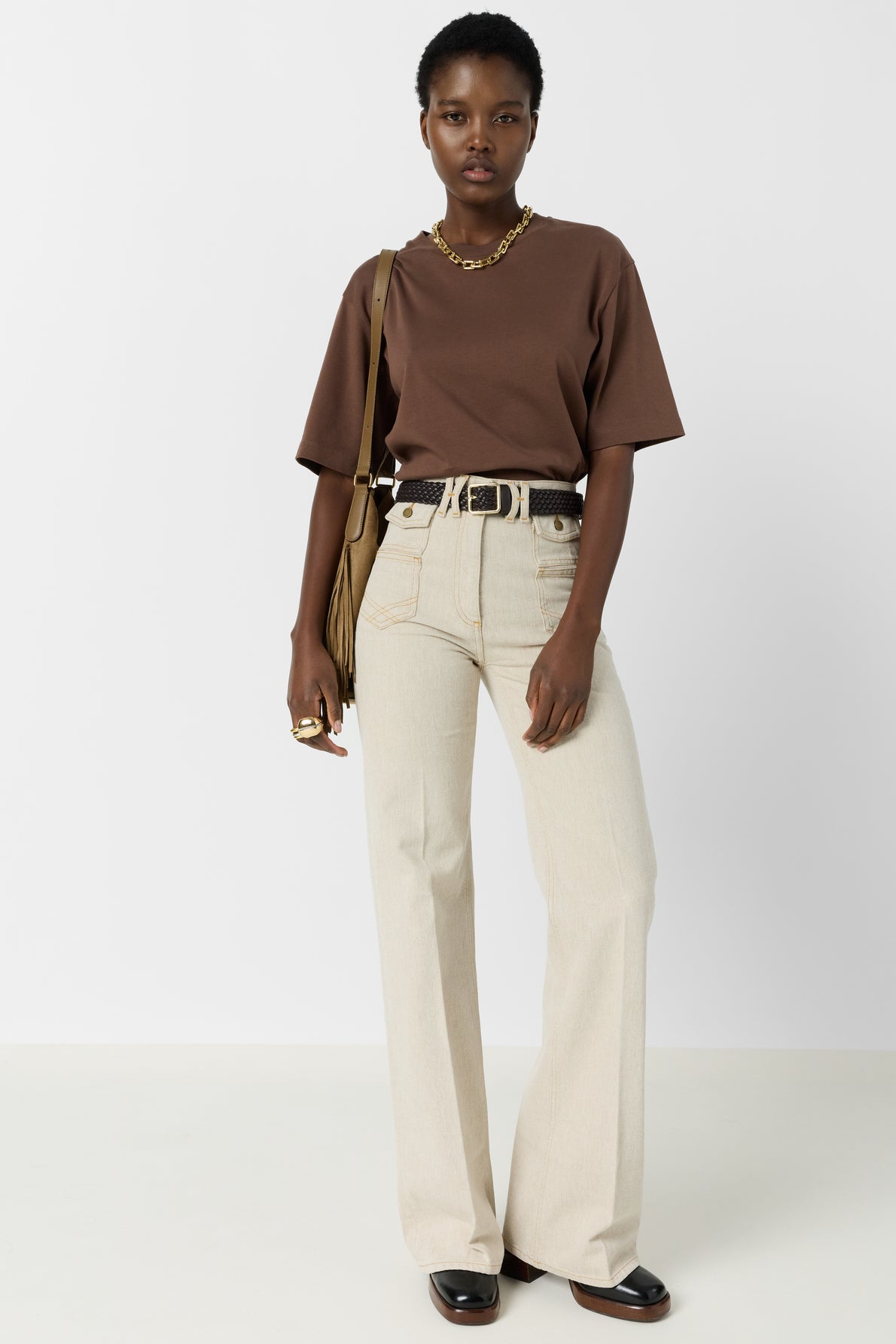 A person stands against a white background wearing a brown t-shirt, beige high-waisted pants, a black belt, dark shoes, a gold necklace, rings, and a brown fringed shoulder bag.