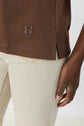 A person wearing light beige jeans and a brown top with a small embossed logo near the hem, standing against a light background, with their right arm visible.