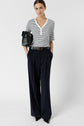 A person wearing a striped short-sleeve shirt, dark wide-leg trousers, black belt, and pointed shoes stands holding a black clutch under one arm, posing against a plain white background.