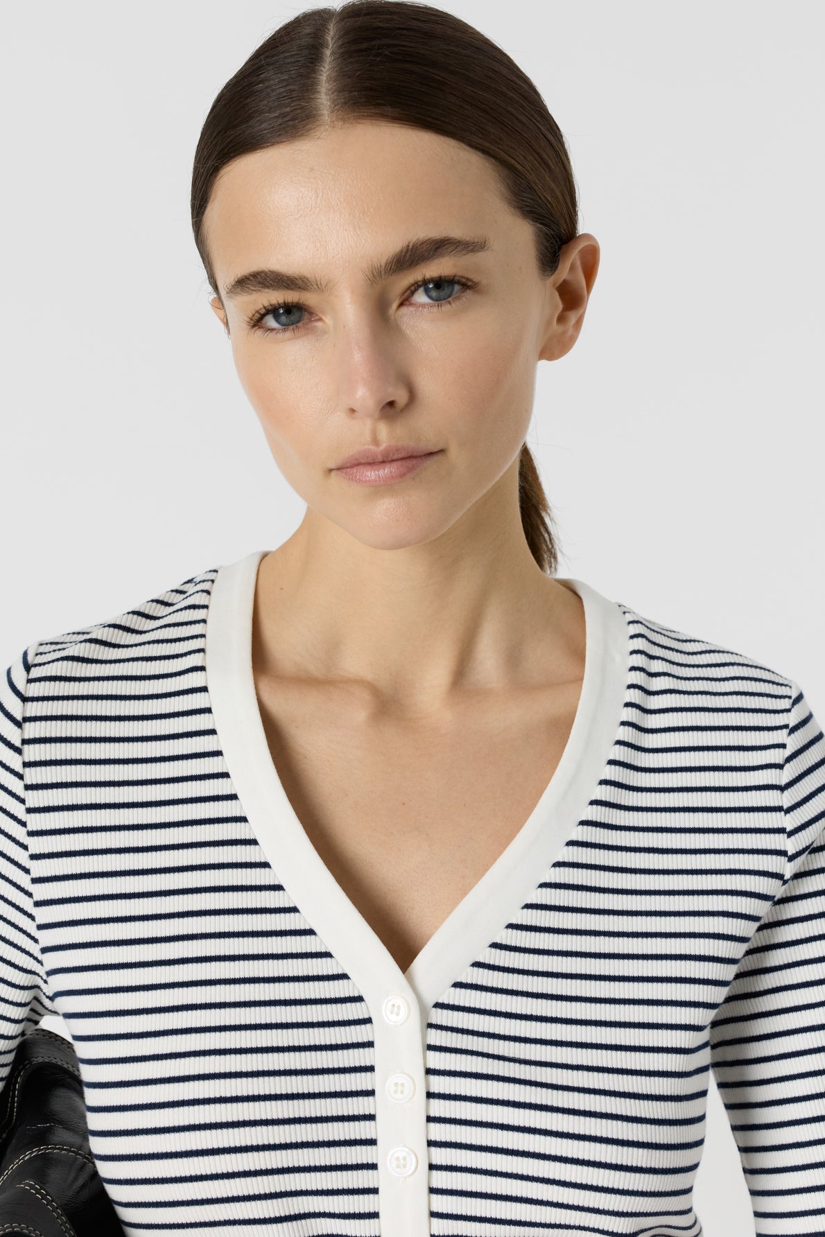 Woman with straight brown hair pulled back, wearing a white and black striped V-neck buttoned shirt, looks directly at the camera with a neutral expression against a plain background.