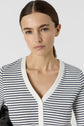Woman with straight brown hair pulled back, wearing a white and black striped V-neck buttoned shirt, looks directly at the camera with a neutral expression against a plain background.
