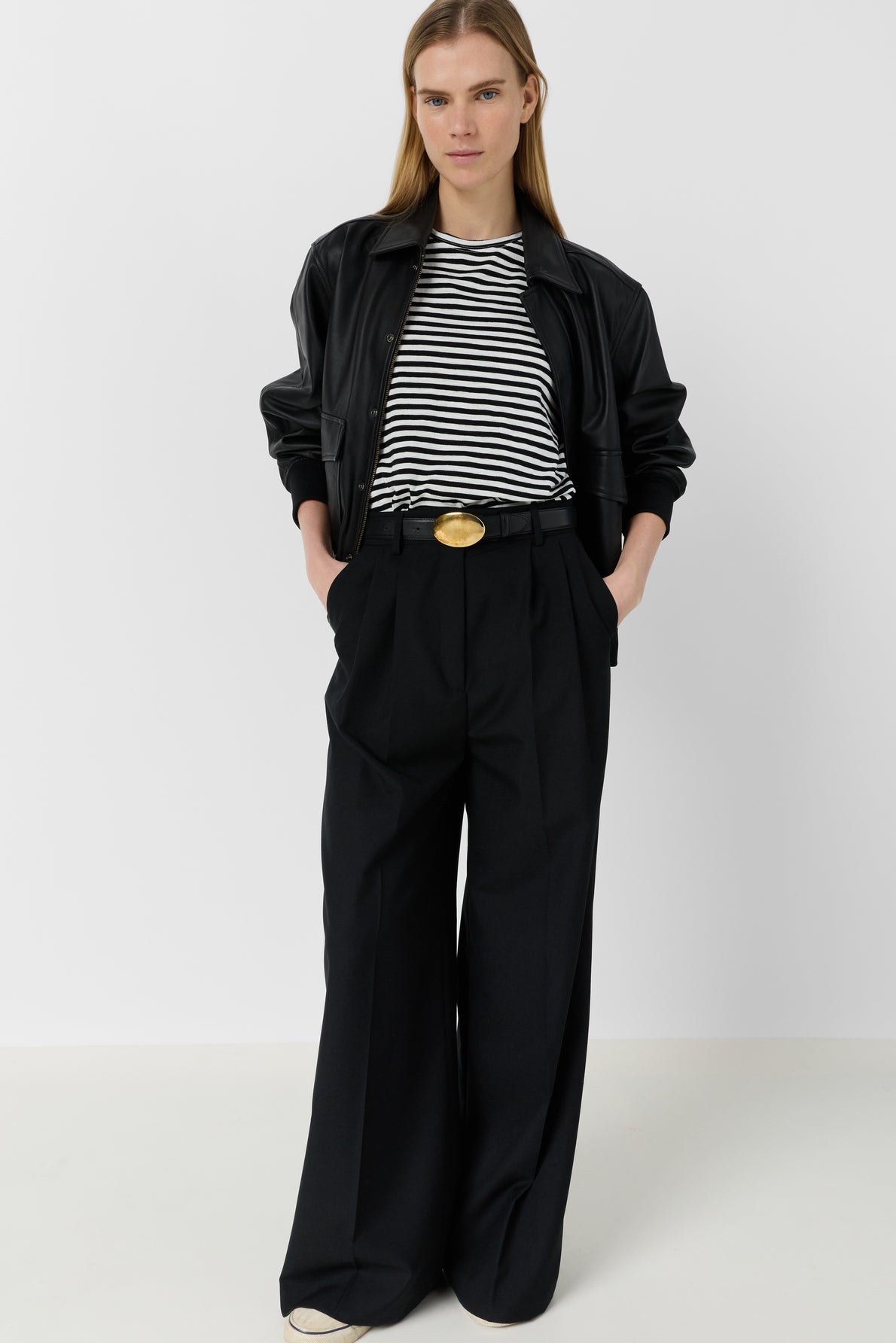 A person wearing a black leather jacket, a tee-shirt Mayane à rayures noires et blanches avec des manches courtes papillon, wide-leg black trousers with a large belt buckle, and white shoes stands against a plain white background.