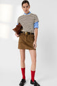 A person wearing a striped t-shirt over a blue shirt, brown mini skirt with a black belt, red knee-high socks, black loafers, and holding a brown clutch bag stands against a plain background.