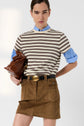 Person wearing a striped short-sleeve shirt over a blue button-up, tan mini skirt with large buckle belt, and holding a brown bag under their arm, standing against a plain light background.