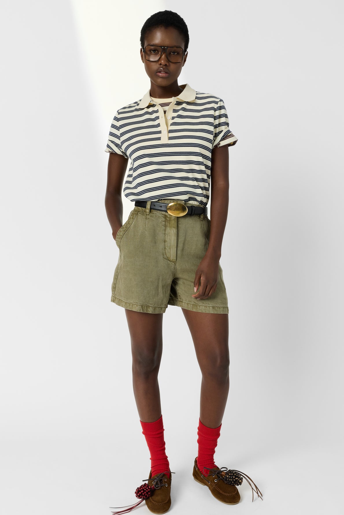 Person wearing a striped short-sleeve polo shirt, olive shorts with a belt, red socks, and brown moccasins with decorative bows, standing with hands in pockets and glasses on. White background.