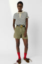 Person wearing a striped short-sleeve polo shirt, olive shorts with a belt, red socks, and brown moccasins with decorative bows, standing with hands in pockets and glasses on. White background.