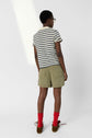 Person standing with back to camera, wearing a striped short-sleeve shirt, olive green shorts, red socks, and brown shoes. The background is plain white.