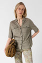 A woman with blonde hair wears a light olive button-up shirt, floral pants, and holds a brown textured handbag. She stands in front of a plain white background, gazing directly at the camera.