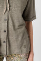 A person wearing an olive green button-up shirt with rolled sleeves and floral patterned pants, shown from the chest down.
