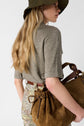 Woman in a green hat and grey shirt with long blonde hair, holding a woven brown bag. She is wearing patterned pants with a brown belt, standing against a plain white background.