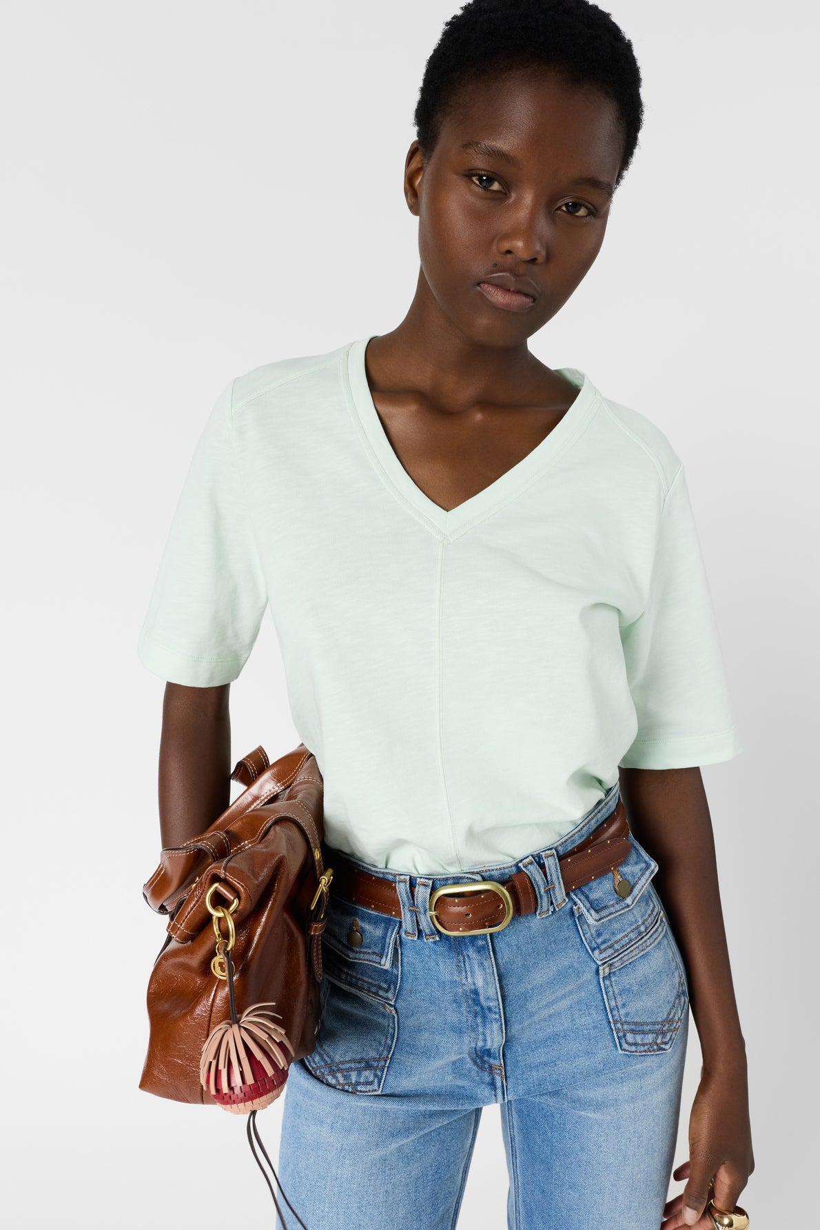 A person wearing a light green V-neck T-shirt and blue jeans holds a brown leather handbag. They have short hair and a neutral expression, standing against a plain white background.