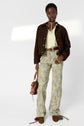 A person stands wearing a brown jacket, light yellow shirt, floral print pants, and brown shoes, holding a brown handbag. The background is plain white with a blurred light shape on the wall.