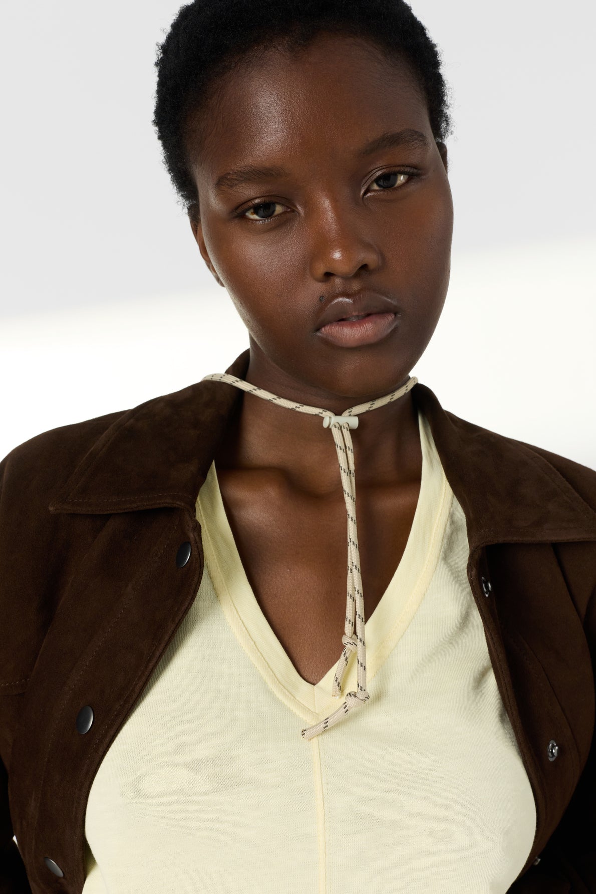 Woman with short curly hair wears a brown jacket over a pale yellow top and a necklace made of thin rope tied in a knot at her neck. She has a neutral facial expression.