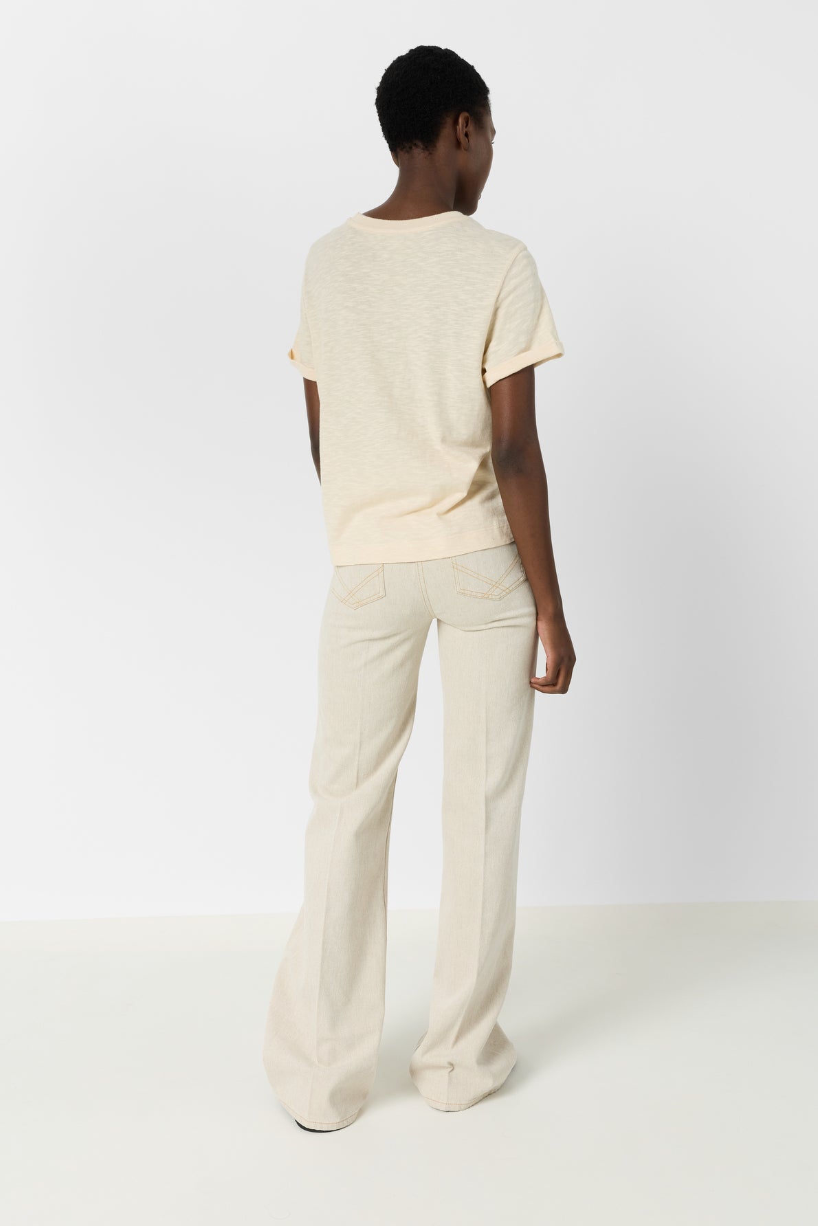 A person stands in a minimal studio, facing away. They wear a short-sleeve cream t-shirt femme and light beige wide-leg pants. The setting is simple with a white wall and floor.