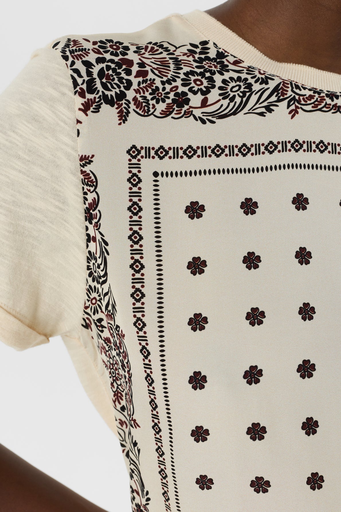 Close-up of a cream-colored t-shirt femme with a black and red floral and geometric border pattern and small t-shirt motif floral details throughout. Part of a person’s shoulder, arm, and neck are visible.
