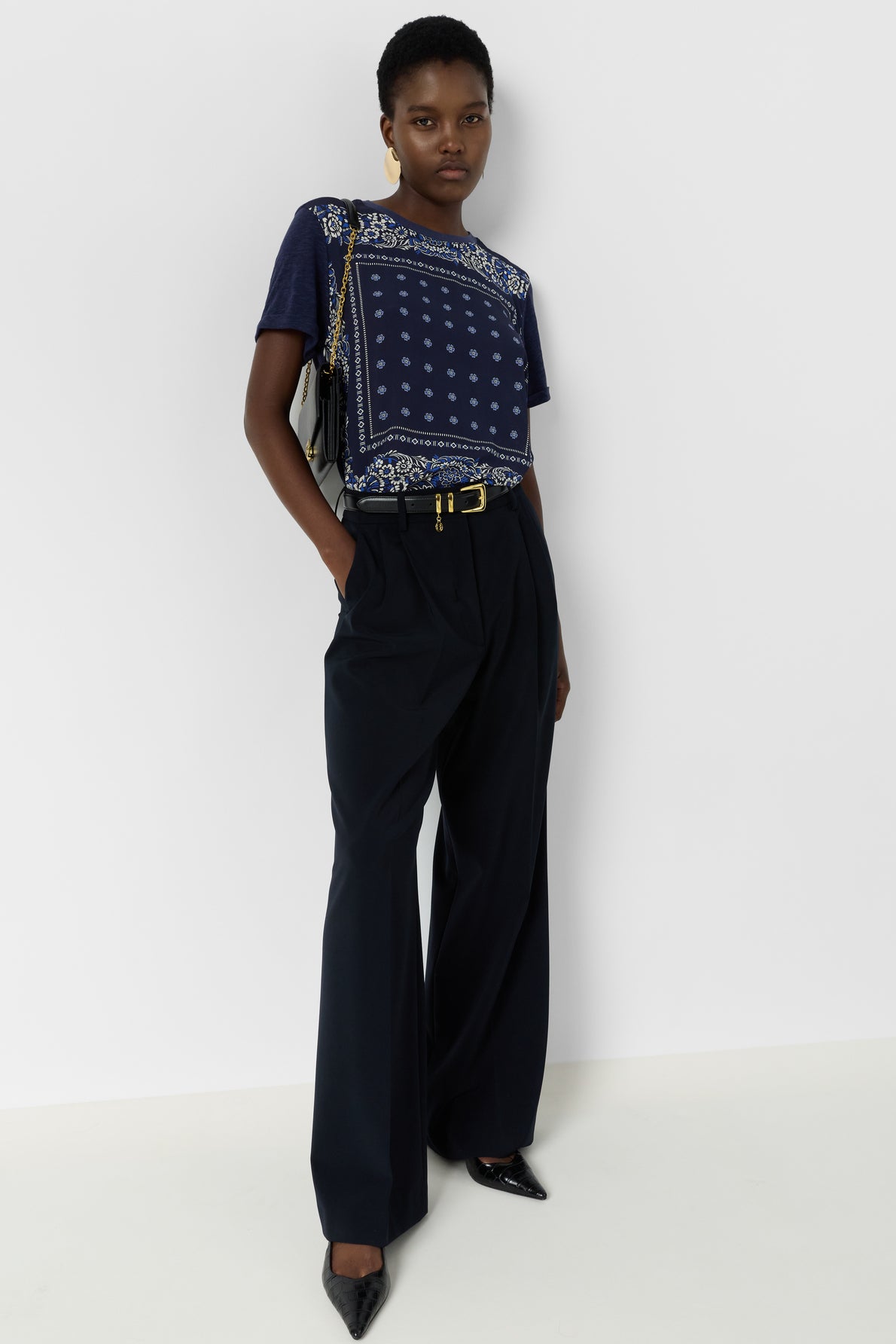 A person stands against a white background wearing a navy printed t-shirt, high-waisted dark wide-leg trousers, black pointed shoes, a belt, and a shoulder bag. Hands are in pockets, expression is neutral.