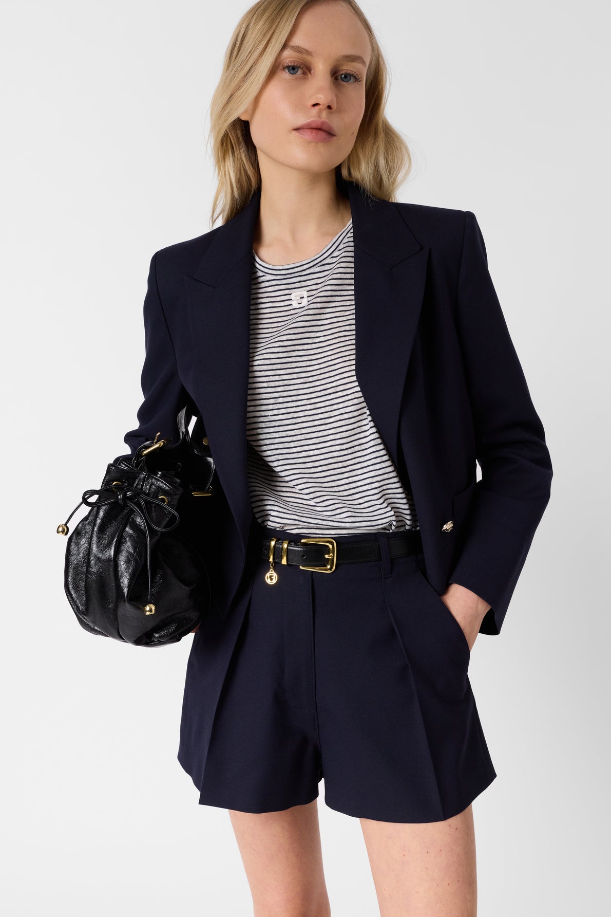 A woman wearing a navy blazer, striped t-shirt, and matching navy shorts holds a black leather drawstring bag. She has blonde hair and stands against a plain white background.