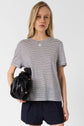 A person with long blonde hair wears a black-and-white striped t-shirt and black shorts, holding a black handbag, standing against a plain light background.
