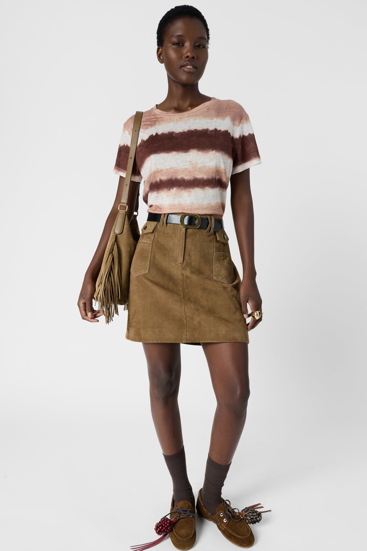 A person wearing a striped tie-dye t-shirt, brown skirt, black belt, brown socks, loafers, and a fringe bag stands against a plain white background.