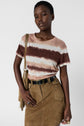 Person wearing a brown and white tie-dye t-shirt, khaki suede skirt with buttons, black belt, and holding a tan fringe bag, standing against a plain white background.