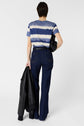 Person standing with back to camera, wearing a blue and white striped t-shirt, high-waisted dark jeans, and holding a black jacket and a purse. The background is plain and white.