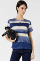 Woman wearing a short-sleeve blue and white tie-dye t-shirt and dark jeans, holding a black bag under her arm, standing against a plain white background.