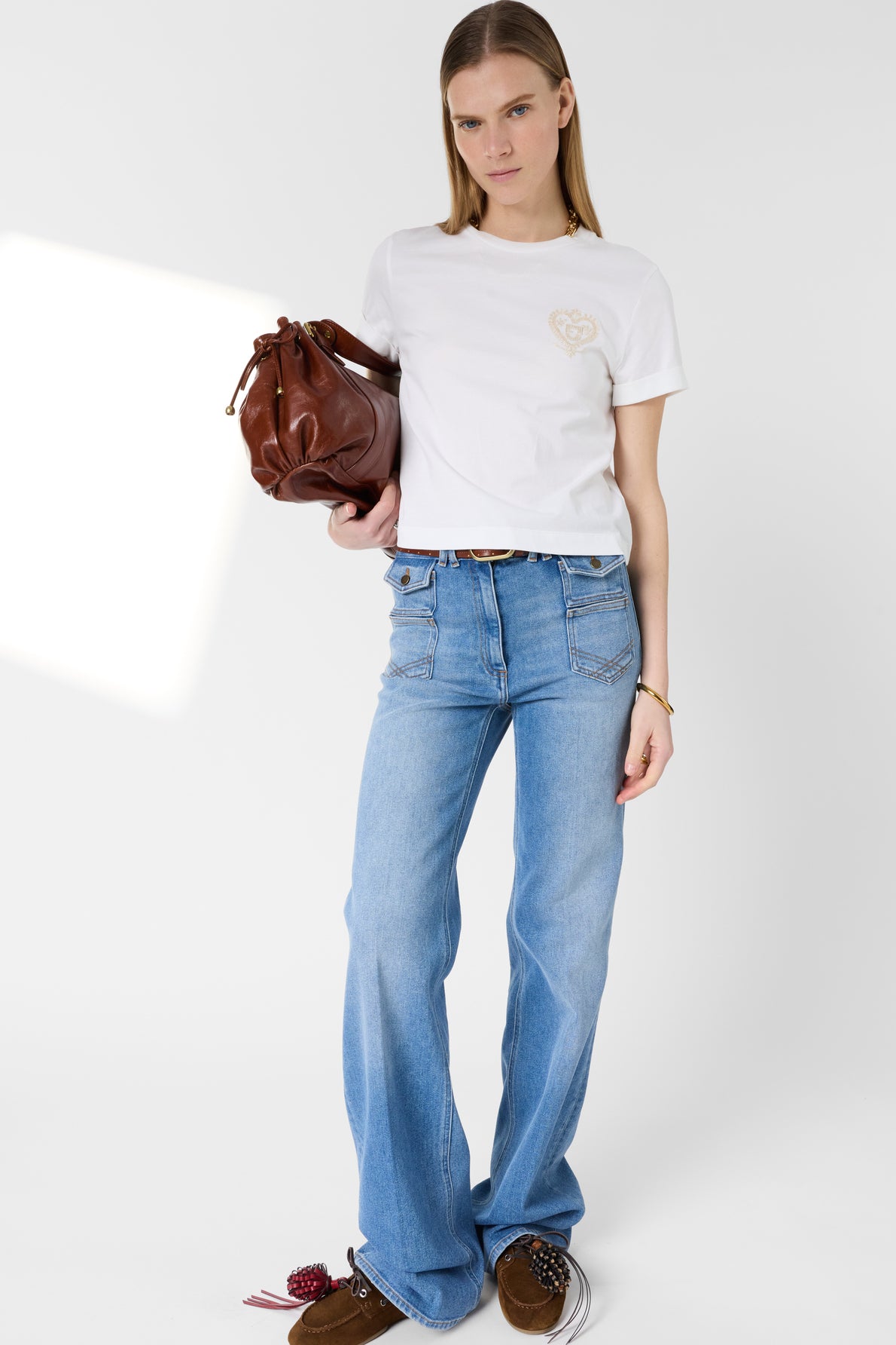 A person wearing a white t-shirt and blue jeans holds a brown handbag. They have straight, light brown hair and are standing against a plain white background.